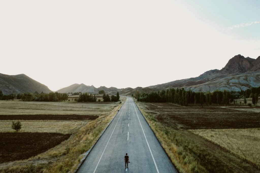 pexels-photo-2902747-2902747-1 A lone person walks on a rural highway surrounded by fields and mountains under a vast sky.